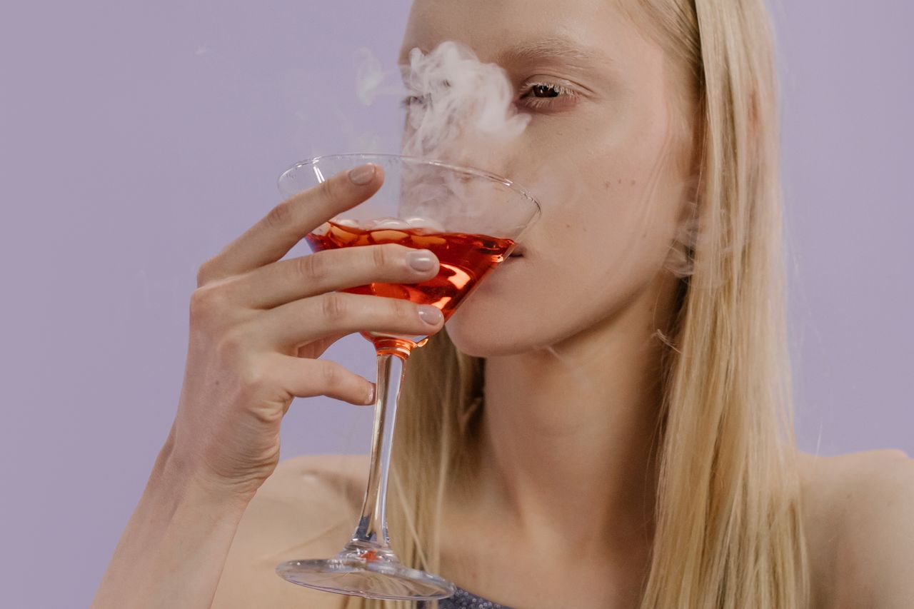 A woman savoring a vibrant, smoky cocktail in a martini glass with blonde hair indoors.